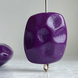 May include: A close-up of two faceted, deep purple beads. One bead is strung on a silver-colored head pin with a gold-colored loop. The other bead is in the background. The beads have a glossy finish and a unique, textured surface.