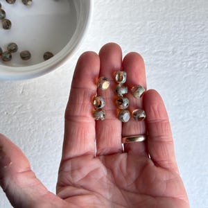 May include: A collection of small, round beads with a mottled, iridescent finish in shades of gray, gold, and blue. Some beads are held in a hand, while others are in a white bowl. The beads have a slightly irregular shape, suggesting they are handmade.