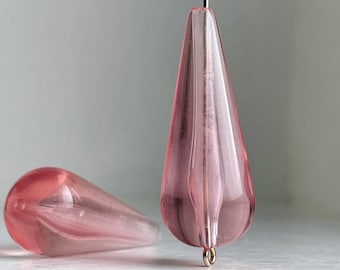 Pode incluir: Duas contas de vidro rosa em forma de lágrima com um furo no topo. A conta à direita tem um laço de metal dourado preso ao furo.