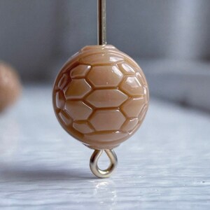May include: A close-up of a beige, round, textured bead with a gold loop. The bead has a honeycomb pattern.