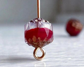 Pode incluir: Close-up de uma conta de vidro vermelha e dourada com um laço dourado preso. A conta é transparente com um centro vermelho e manchas douradas.