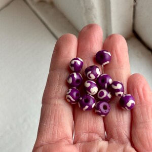 May include: A collection of purple beads with white swirl patterns. The beads are round and have a glossy finish. They are held in a hand, showcasing their size and design. These beads are suitable for jewelry making and crafts.