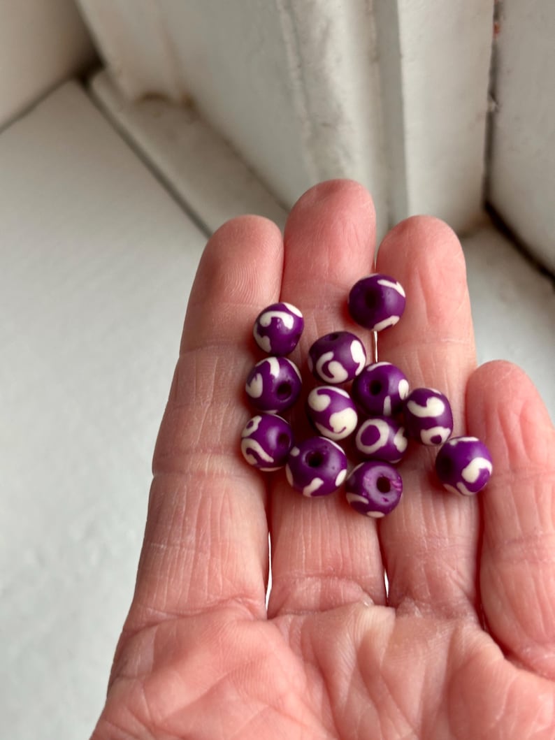 May include: A collection of purple beads with white swirl patterns. The beads are round and have a glossy finish. They are held in a hand, showcasing their size and design. These beads are suitable for jewelry making and crafts.