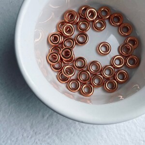 May include: A white bowl filled with copper-colored metal jump rings. The rings are all the same size and are arranged in a random pattern.