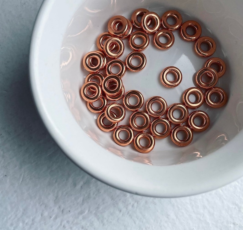 May include: A white bowl filled with copper-colored metal jump rings. The rings are all the same size and are arranged in a random pattern.