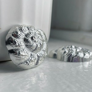 Pode incluir: Dois enfeites decorativos de metal prateado em forma de concha. Os enfeites têm uma superfície texturizada e detalhada.