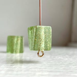 May include: A pair of green glass beads with gold glitter accents. The beads are square shaped and have a small gold loop attached to the top.