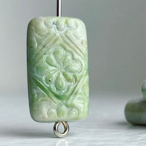 Puede incluir: Una cuenta rectangular verde claro con un diseño floral tallado. La cuenta tiene un pasador de ojo plateado en la parte inferior y está ensartada en un alambre plateado. La cuenta es de color verde claro con acentos verdes más oscuros.