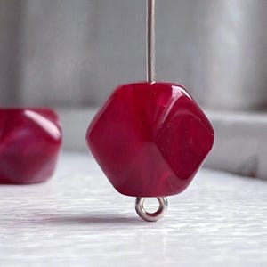 May include: A close-up of a deep red, faceted glass bead with a silver loop attached. The bead is on a white surface.