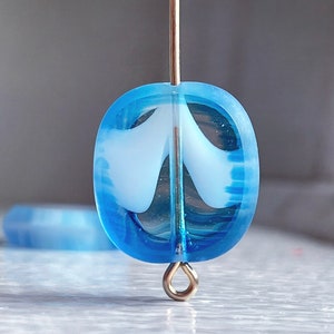 May include: A translucent blue glass bead with a white swirl design. The bead is oval-shaped and has a gold-colored metal eye pin. Another bead is visible in the background.