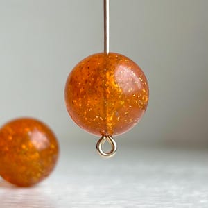 May include: Close-up of a round, orange bead with gold flecks, suspended by a thin wire. The bead has a loop at the bottom. Another similar bead is visible in the background. Jewellery making supplies.