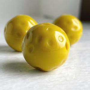 Pode incluir: Três contas de vidro amarelo com uma superfície texturizada. As contas são redondas e têm um acabamento brilhante.