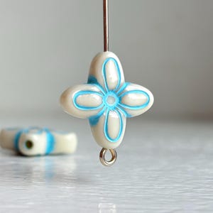 May include: A close-up of a white and blue flower-shaped bead. The bead is strung on a silver-colored head pin. Another bead is out of focus in the background. The bead has a four-petal design with blue accents.