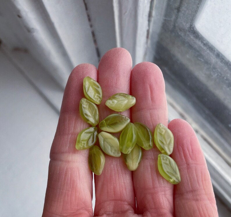 Op de afbeelding: Een verzameling groene glazen kralen in de vorm van bladeren. De kralen zijn doorschijnend en hebben een licht gematteerde afwerking.