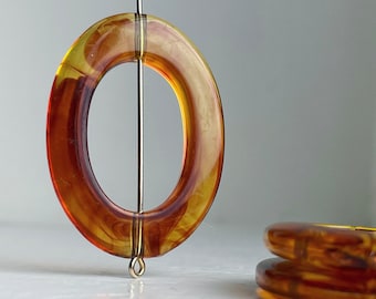 Op de afbeelding: Een grote, ovale ring van amberkleur met een swirl-patroon. De ring hangt aan een dunne metalen draad.