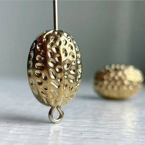 Könnte beinhalten: Ein goldfarbener Metallohrring mit einer strukturierten, unebenen Oberfläche. Der Ohrring ist oval geformt und hat oben eine kleine Schlaufe zum Anbringen an einen Haken.