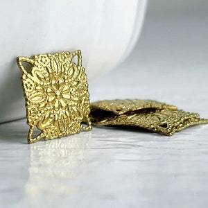 May include: Gold metal square embellishments with a detailed floral design. The embellishments are stacked on top of each other.