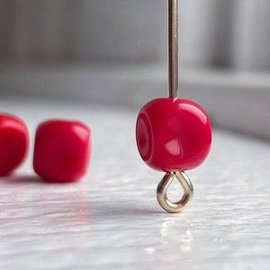 May include: Close-up of a bright red, cube-shaped bead with rounded edges, strung on a gold-colored wire. The bead has a glossy finish and is suspended above a gold-colored loop. Two additional red beads are slightly out of focus in the background.