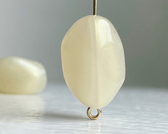 Pode incluir: Uma gema oval branca com um laço dourado preso. A gema tem um brilho perolado.