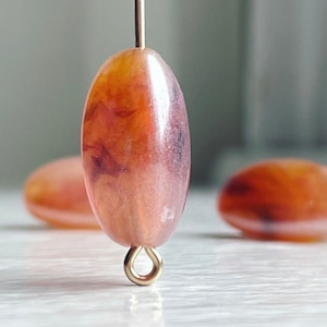 May include: A close-up of a single, oval-shaped, orange and brown glass bead with a gold metal loop attached. The bead has a marbled, swirled pattern.