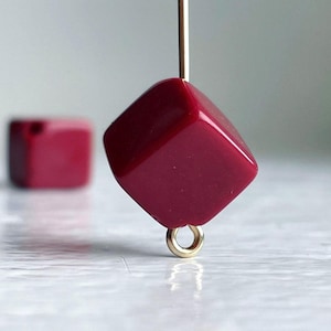 May include: A deep red square bead with a gold loop attached. The bead is hanging from a gold wire.