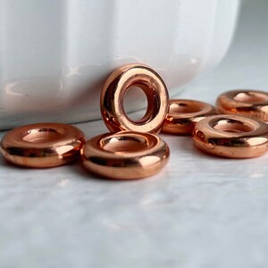 May include: Six shiny copper-colored metal rings, each with a smooth, rounded surface and a hole in the center. The rings are arranged in a semi-circle on a white surface.