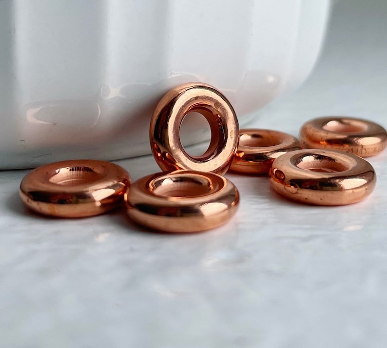 May include: Six shiny copper-colored metal rings, each with a smooth, rounded surface and a hole in the center. The rings are arranged in a semi-circle on a white surface.