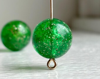Puede incluir: Primer plano de una cuenta verde esférica con motas doradas, suspendida por un alambre dorado. Otra cuenta similar está ligeramente desenfocada en el fondo. La cuenta tiene un lazo en la parte inferior para sujetar.