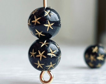 May include: Two black beads with gold star designs are strung on a gold wire. The beads are round and have a glossy finish.