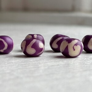 May include: Six purple beads with a white swirl design. The beads are round and have a smooth surface. The beads are likely used for jewelry making or crafts.