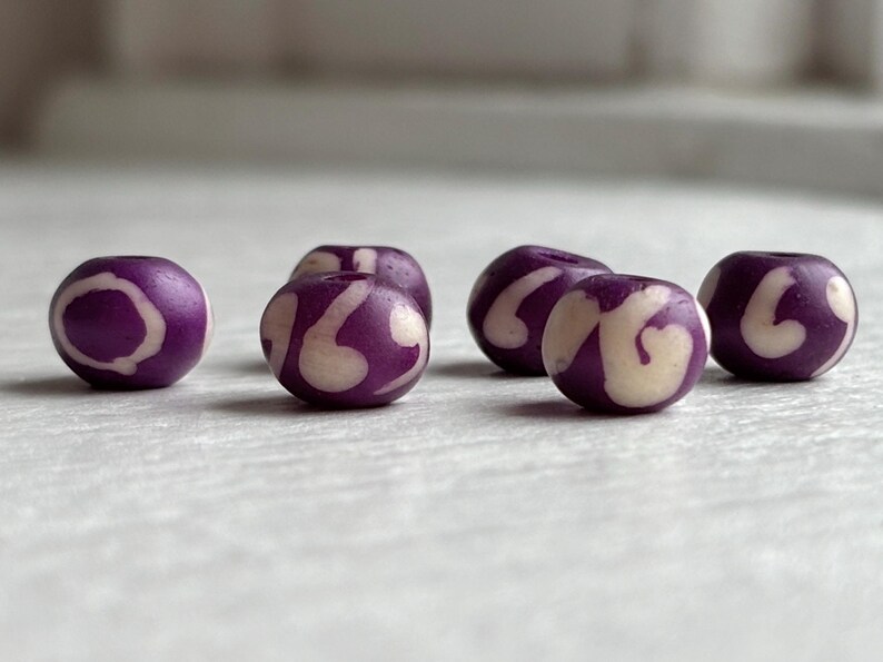 May include: Six purple beads with a white swirl design. The beads are round and have a smooth surface. The beads are likely used for jewelry making or crafts.