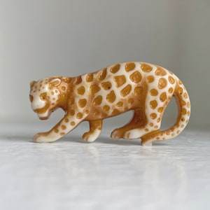May include: A ceramic figurine of a leopard with brown and white spots. The figurine is in a crouching position, with its head turned to the side.