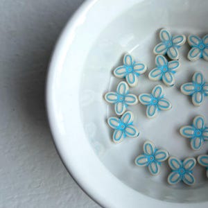 May include: A white ceramic bowl filled with small, flower-shaped beads. The beads are white with a light blue outline and center, creating a delicate floral design. The bowl sits on a white surface.