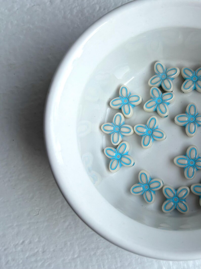 May include: A white ceramic bowl filled with small, flower-shaped beads. The beads are white with a light blue outline and center, creating a delicate floral design. The bowl sits on a white surface.