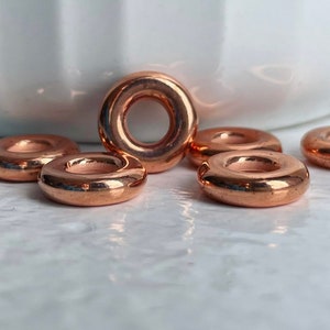 May include: Six copper-colored metal rings, each with a smooth, rounded surface and a hole in the center. The rings are arranged in a semi-circle on a white marble surface.