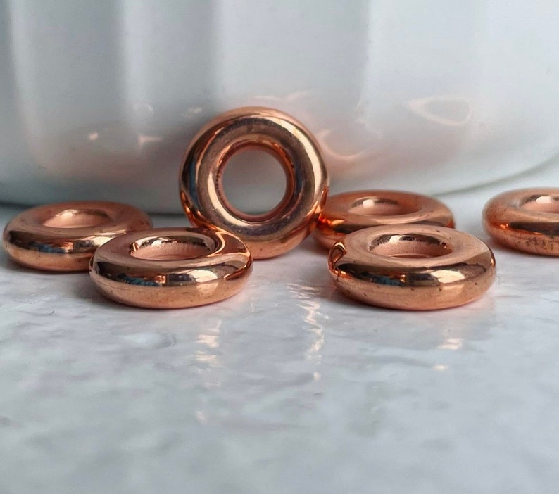 May include: Six copper-colored metal rings, each with a smooth, rounded surface and a hole in the center. The rings are arranged in a semi-circle on a white marble surface.
