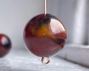 Può includere: Primo piano di una perla rotonda e lucida con un mix di colori bordeaux, giallo e nero. La perla è sospesa da un sottile filo color rame e ha un piccolo anello dorato nella parte inferiore, suggerendo che sia per la creazione di gioielli.