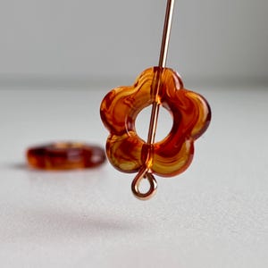 May include: Close-up of a flower-shaped bead with a hole in the center, strung on a copper wire. The bead is a translucent amber color with swirling patterns. Another similar bead is out of focus in the background.
