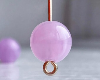 Pode incluir: Uma única conta de vidro redonda de cor rosa claro com um laço prateado preso na parte inferior. A conta está pendurada em um fio fino.