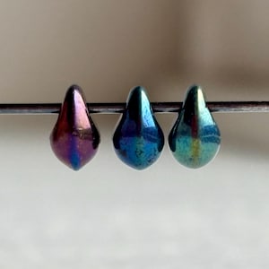 May include: Three iridescent teardrop-shaped beads in shades of purple, blue, and green are displayed on a thin metal rod. The beads have a glossy finish and reflect light, creating a shimmering effect. These beads are suitable for jewelry making.