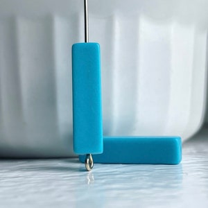 Puede incluir: Dos cuentas rectangulares de arcilla polimérica azul claro con un anillo de salto plateado unido a una cuenta.