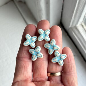 May include: Six small, white flower-shaped beads with light blue accents are displayed. The beads are arranged on a person's hand, with a gold ring visible on one finger. The flowers have a four-petal design.