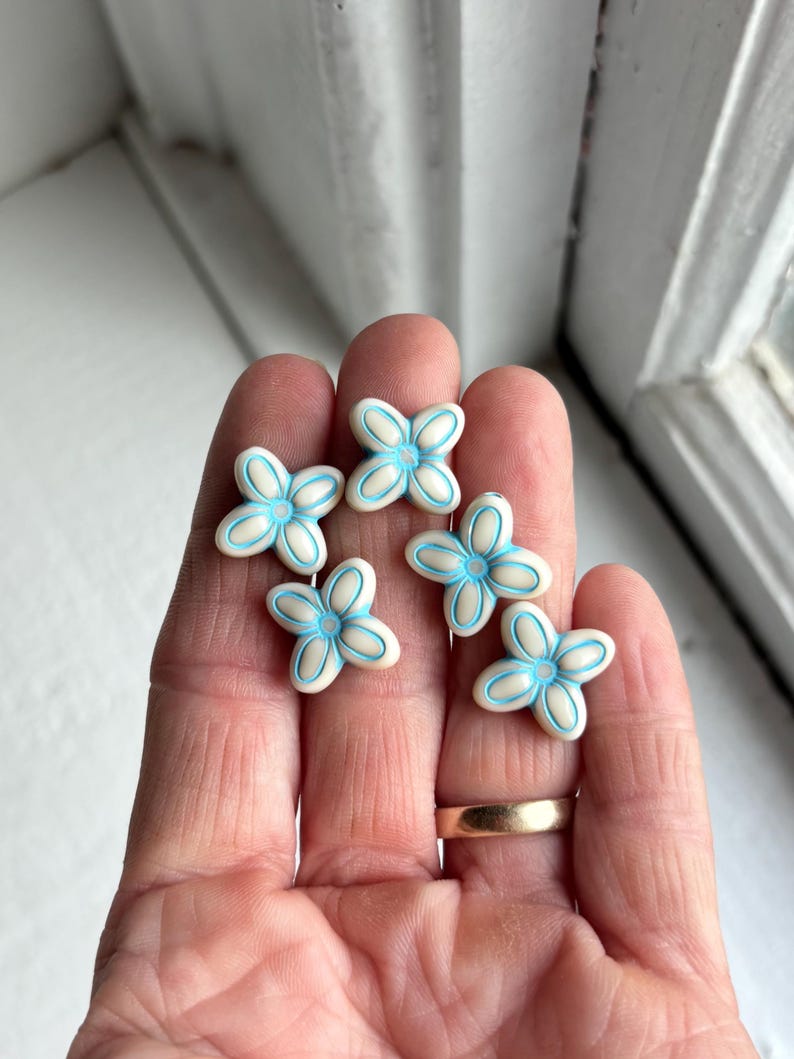 May include: Six small, white flower-shaped beads with light blue accents are displayed. The beads are arranged on a person's hand, with a gold ring visible on one finger. The flowers have a four-petal design.