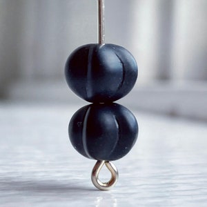 May include: Close-up of two black, spherical beads with vertical grooves, strung on a silver-colored wire. The beads are stacked, with a silver loop at the bottom. The background is blurred, suggesting a focus on the jewelry components.