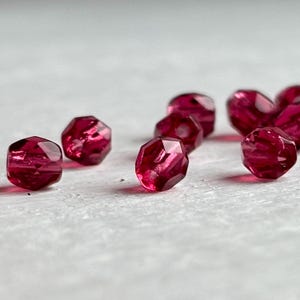 Op de afbeelding: Close-up van verschillende gefacetteerde, dieprode kralen. De kralen hebben een geometrische vorm met meerdere zijden, die het licht reflecteren. Ze zijn verspreid over een lichtgekleurd oppervlak, wat hun kleur en vorm benadrukt.