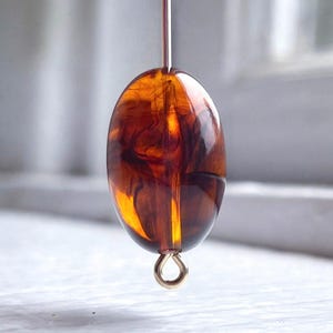 May include: A translucent, oval-shaped bead with a rich amber and brown color gradient. The bead is suspended by a thin, silver-colored wire and a small gold-colored loop at the bottom. The bead's surface reflects light, showcasing its internal patterns.