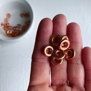 May include: A close-up of a hand holding a pile of small, copper-colored metal rings. The rings are all the same size and shape, and they are arranged in a loose pile.