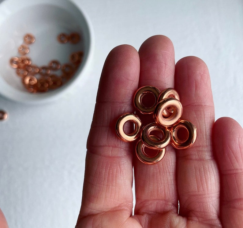 May include: A close-up of a hand holding a pile of small, copper-colored metal rings. The rings are all the same size and shape, and they are arranged in a loose pile.