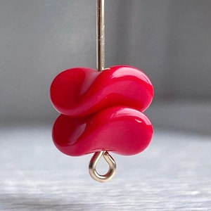 May include: A bright pink glass bead with a unique, twisted shape. The bead is strung on a silver wire and has a gold loop at the bottom.