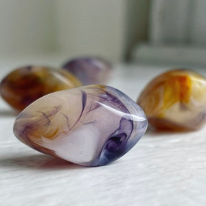 May include: Four abstract swirl patterned glass marbles in shades of purple, white, and brown. The marbles are smooth and glossy.